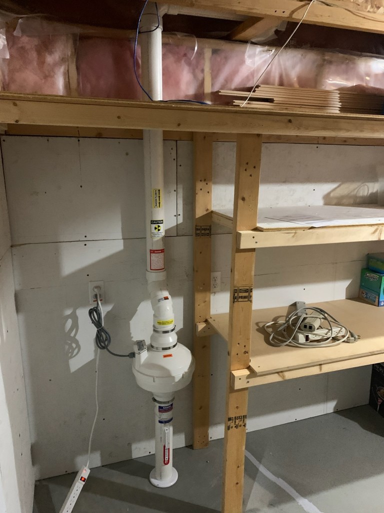 Radon mitigation system installed in a basement, with pipes running vertically along a wall next to wooden shelves.