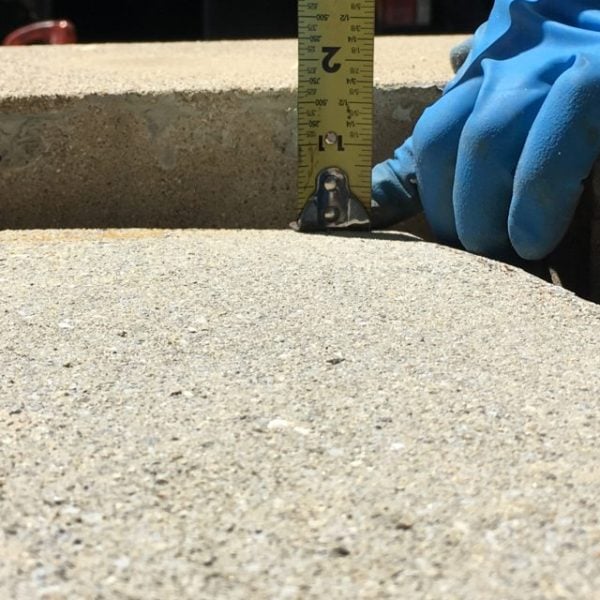 A gloved hand measuring the depth of an opening in a concrete surface with a yellow measuring tape.