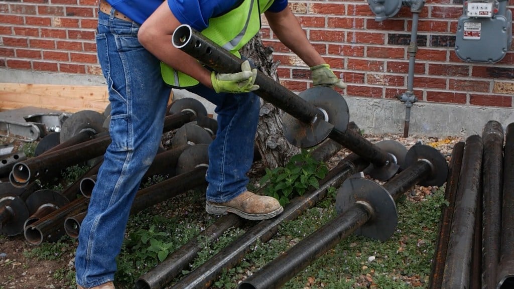 Worker in safety gear handling metal pipes on grass near a brick wall.