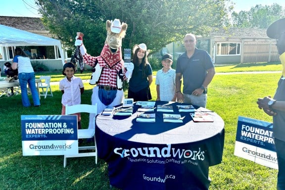 People and a mascot stand by a table with brochures at an outdoor event for Groundworks, a foundation expert company.