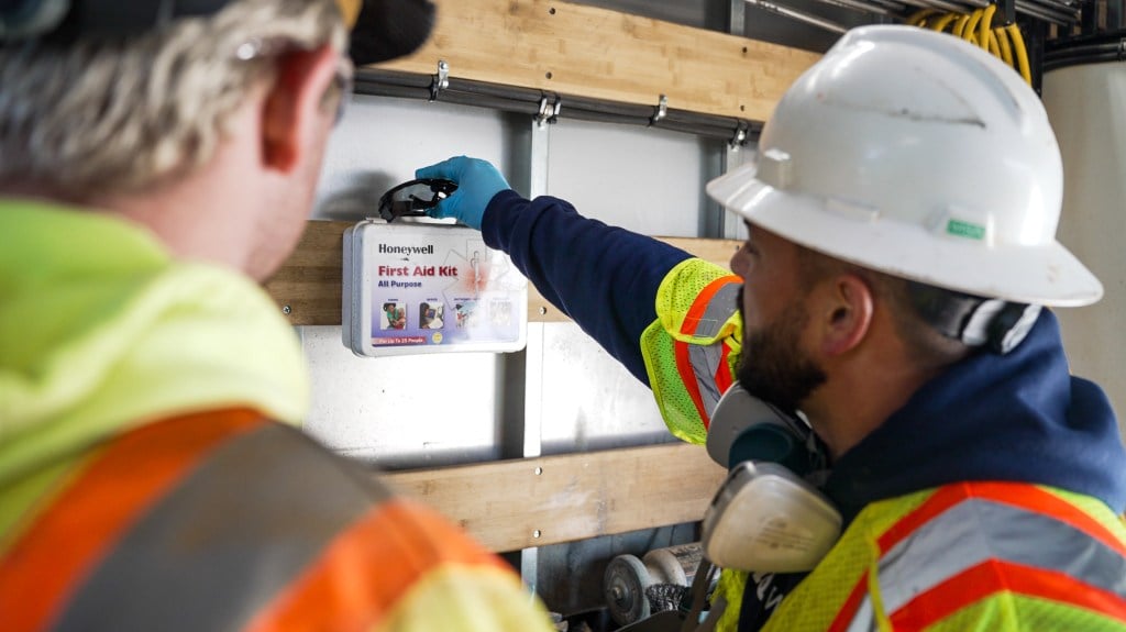 Two construction workers in safety gear check a first aid kit on a wall.