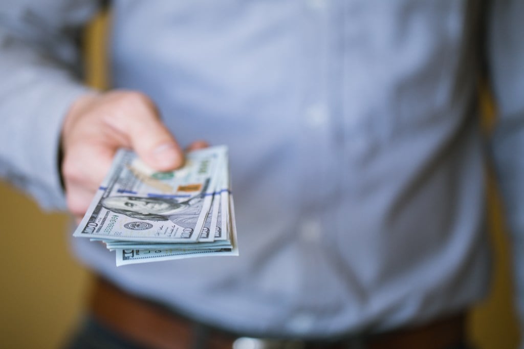 A person in a blue shirt holding a stack of US hundred-dollar bills, extended towards the viewer.